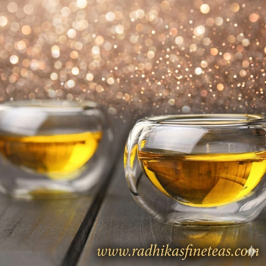 Two clear glass cups filled with tea on a wooden surface with a sparkling background, featuring the brand 'www.radhikasfine teas.