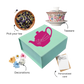 A green box of tea bliss with a pink and white teapot design, accompanied by a white teapot, a small blue flower pot with a purple flower, a decorative card, and a postcard.