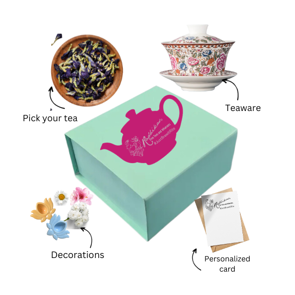 A green box of tea bliss with a pink and white teapot design, accompanied by a white teapot, a small blue flower pot with a purple flower, a decorative card, and a postcard.