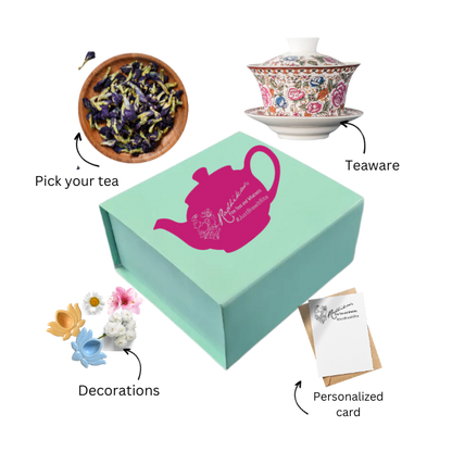 A green box of tea bliss with a pink and white teapot design, accompanied by a white teapot, a small blue flower pot with a purple flower, a decorative card, and a postcard.