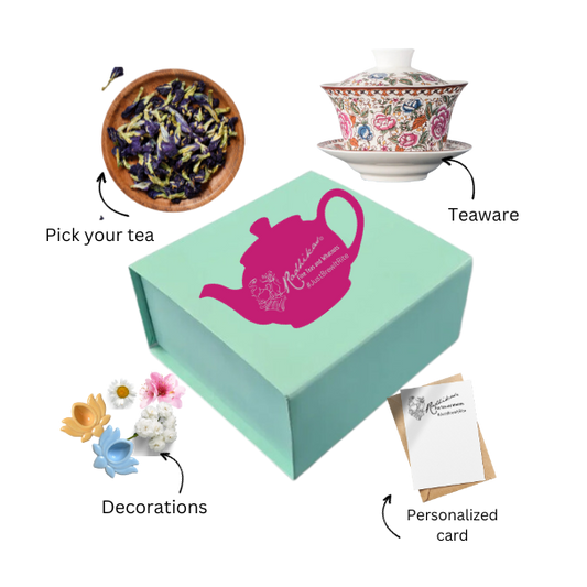 A green box of tea bliss with a pink and white teapot design, accompanied by a white teapot, a small blue flower pot with a purple flower, a decorative card, and a postcard.
