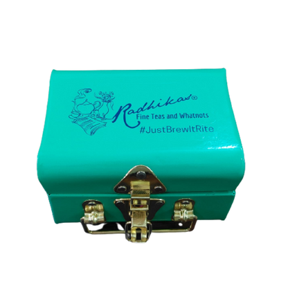 A blue metal tea trunk with the brand name 'Radhikas Fine Teas and Whatnots' and the hashtag 'Just Brew It' on the lid. It has a shiny metal clasp on the front.