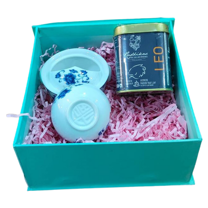 A gift box containing a white ceramic Gaiwan with a blue floral pattern, a small container of tea, and pink tissue paper.