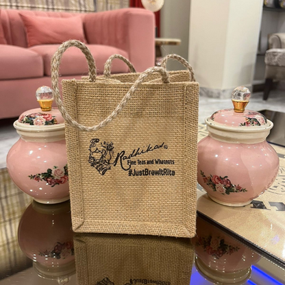 A beige jute tea bag with a handle, branded with the name 'Radhikas Fine Teas and Whatnots', placed on a table with two pink-flowered containers in the background.