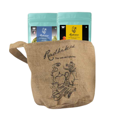 Two ziplocked pouches of Radhikas Fine Teas, one with a matcha green label and the other with a butterfly blue label, placed inside a beige jute bag with an illustration of a tea pot and leaves.