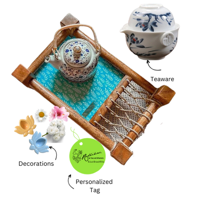 A set of tea equipment including a teapot, a cup, a kettle, and a spoon, displayed on a miniature matte green cloth, with decorative flowers and a branded label in the foreground.