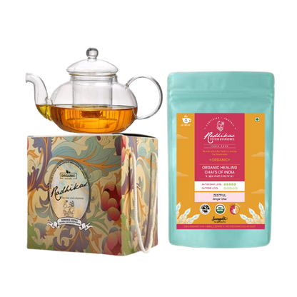 A Victorian glass kettle with a lid next to a packaged tea product, all presented on a decorative box with floral designs.