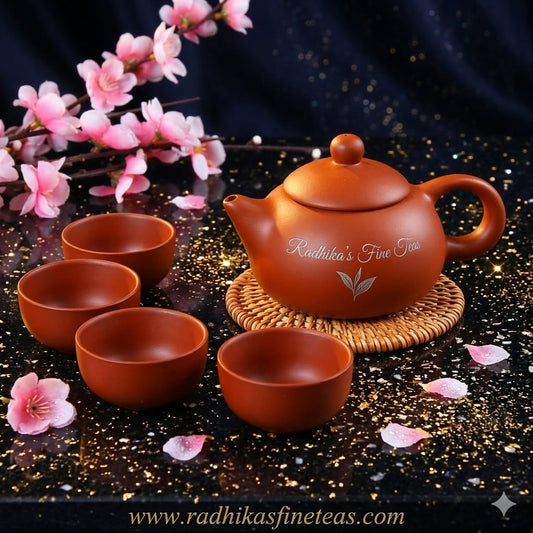 Ceramic teapot and cups with cherry blossoms on a dark surface, featuring Radhika's Fine Teas branding.